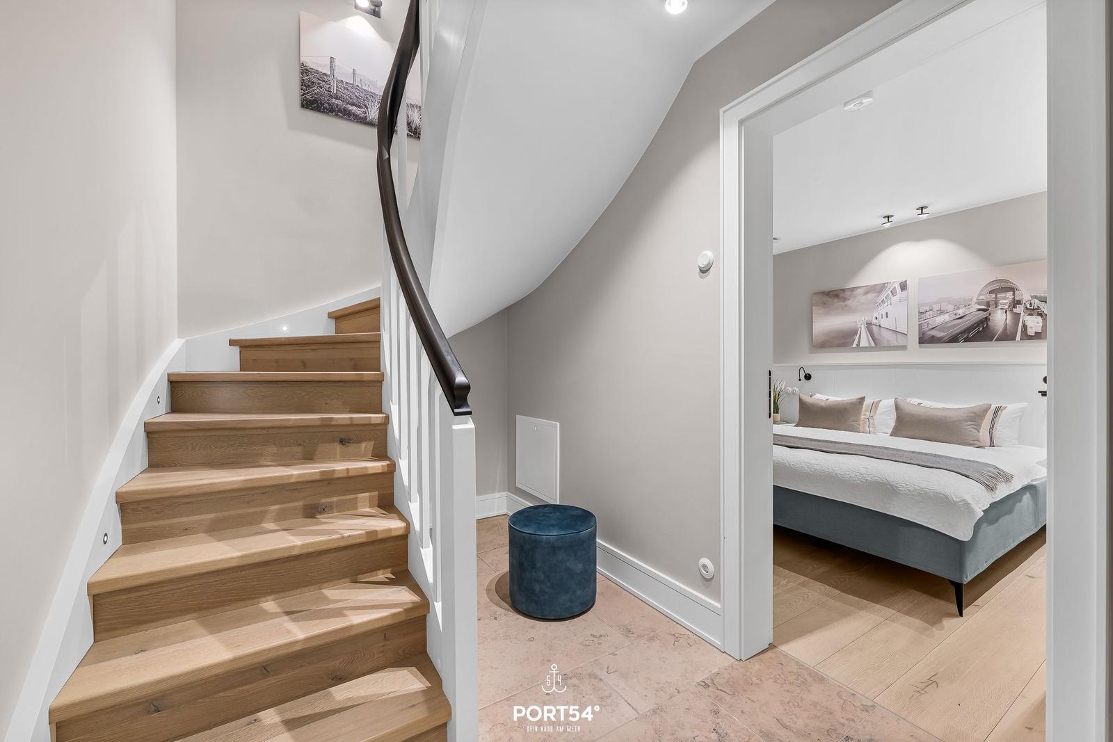 Treppenhaus mit Holztreppe und Blick in Schlafzimmer mit Bett und Wandbildern.