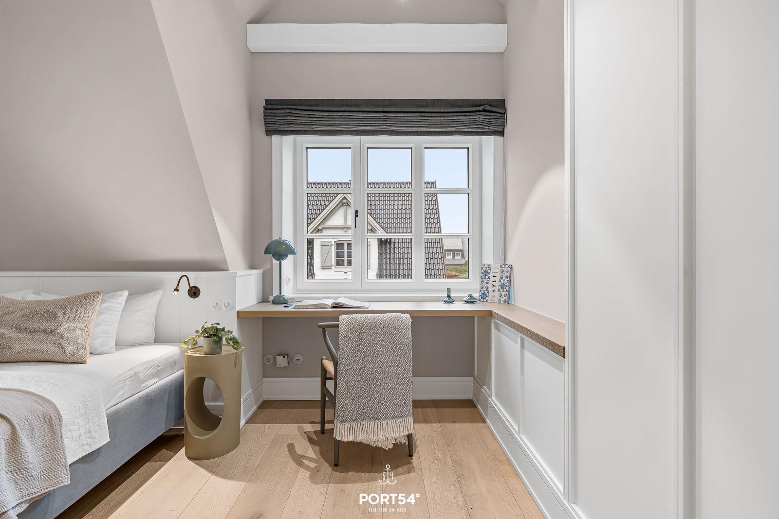 Schlafzimmer mit Schreibtisch und Fensterblick auf Dächer.