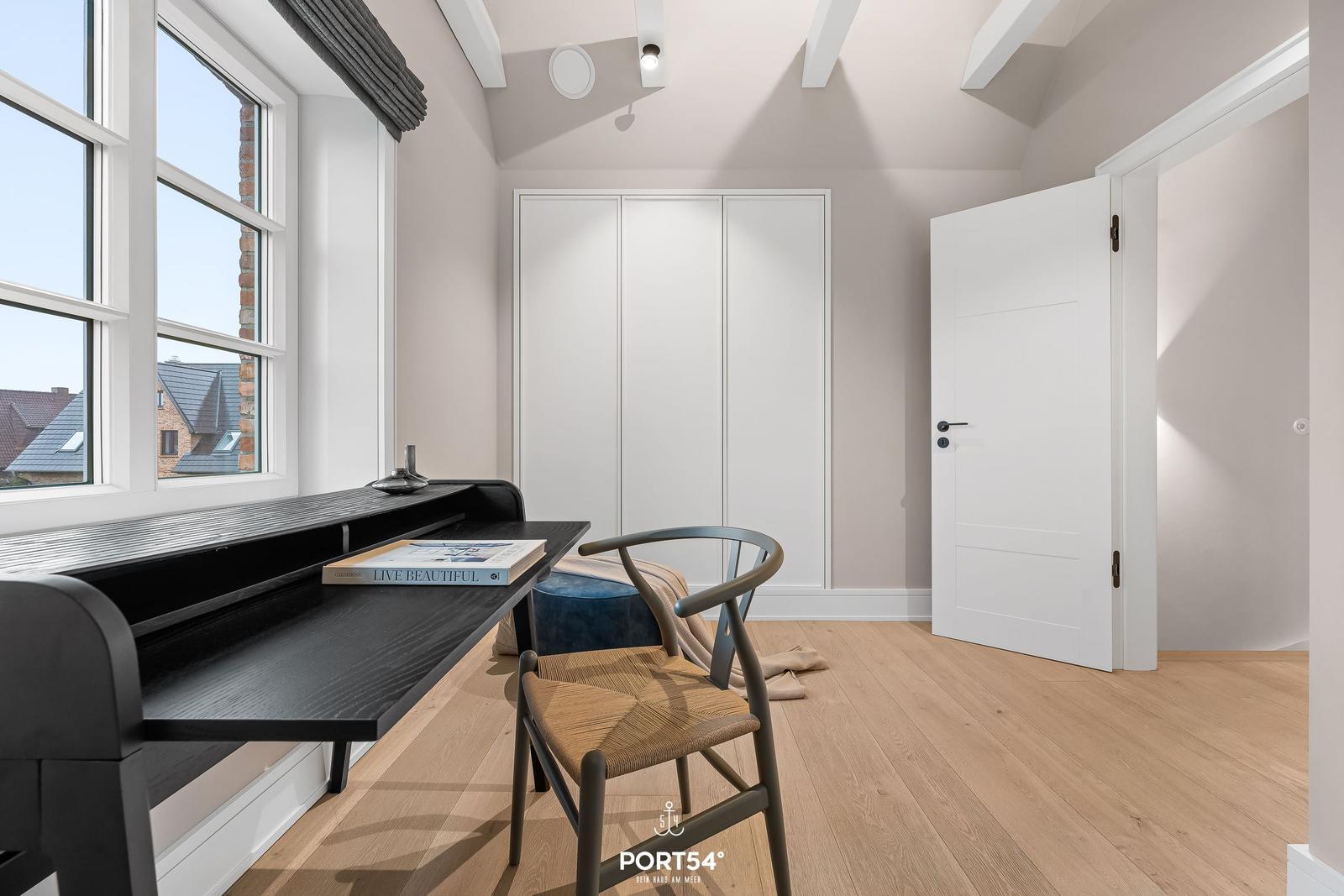 Schlafzimmer mit Schreibtisch, Stuhl und Kleiderschrank. Holzboden und großes Fenster.