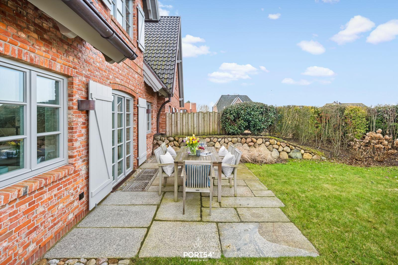 Roter Backsteinbau mit Terrasse und Garten. Holztisch mit Stühlen und Blumen.