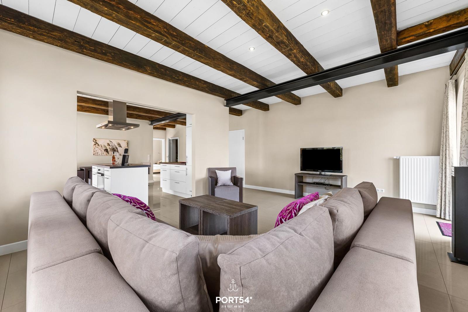Wohnbereich mit Sofa, TV und Küchenbereich. Holzbalken an der Decke.