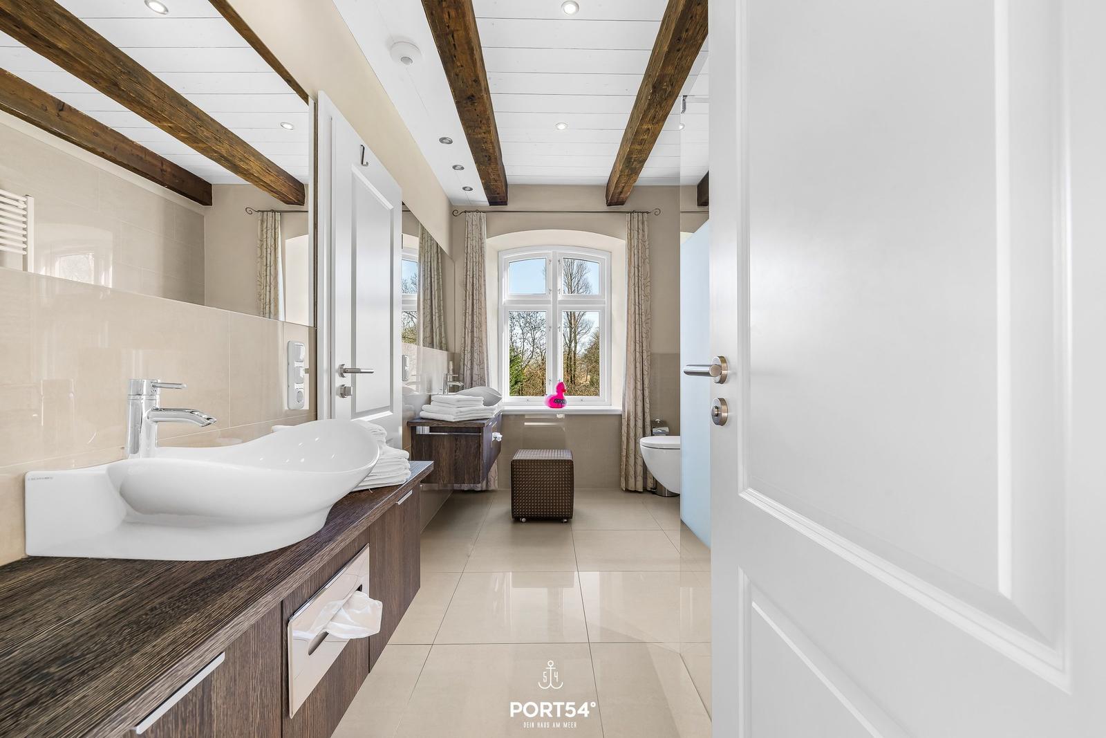 Badezimmer mit Waschbecken, Fenster und Toilette. Holzbalken an der Decke.