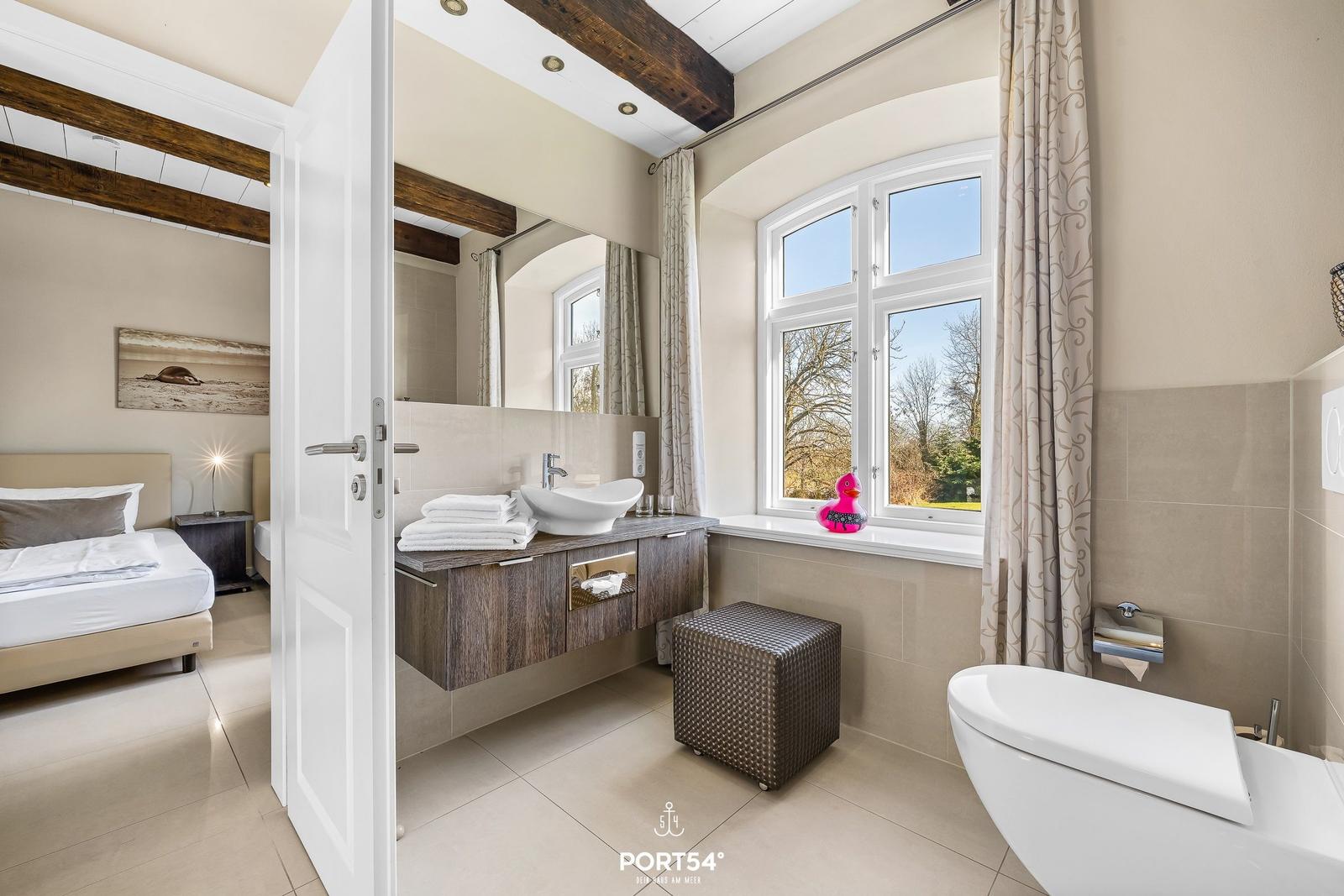 Badezimmer mit Waschbecken, Toilette und Fenster. Schlafzimmer mit Bett und Wandbild sichtbar.