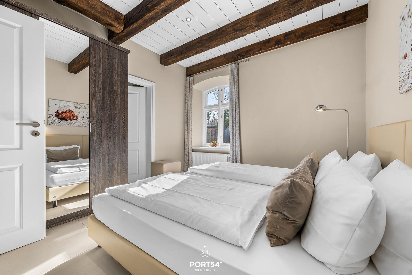 Schlafzimmer mit Doppelbett, Holzbalken und Fenster. Spiegel reflektiert ein weiteres Bett.