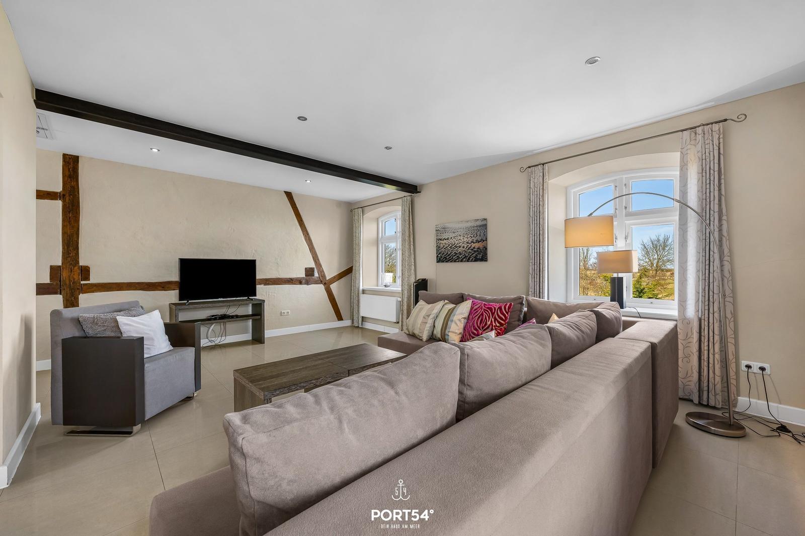 Wohnzimmer mit Sofa, Sessel, Fernseher und großen Fenstern.