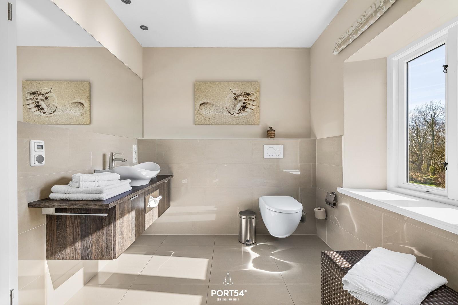 Badezimmer mit Waschtisch, Toilette und Fenster. Wandbilder und Handtuchstapel sichtbar.