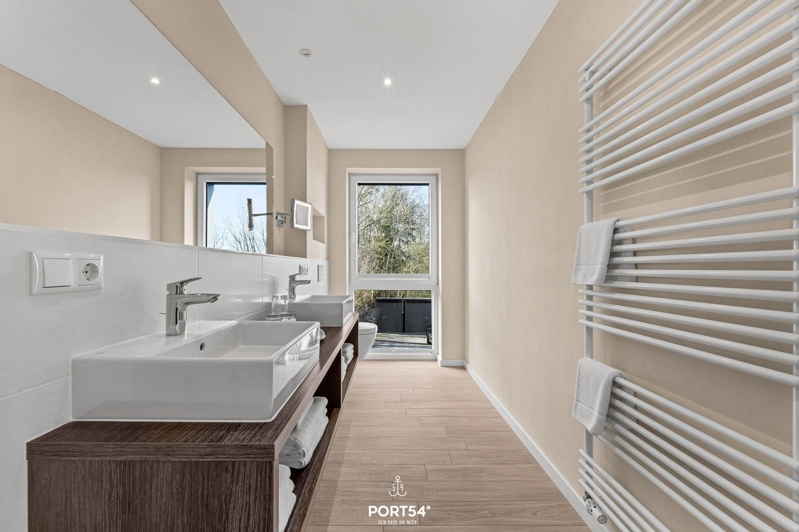 Badezimmer mit Doppelwaschbecken, Heizkörper und Fensterblick auf Außenbereich.