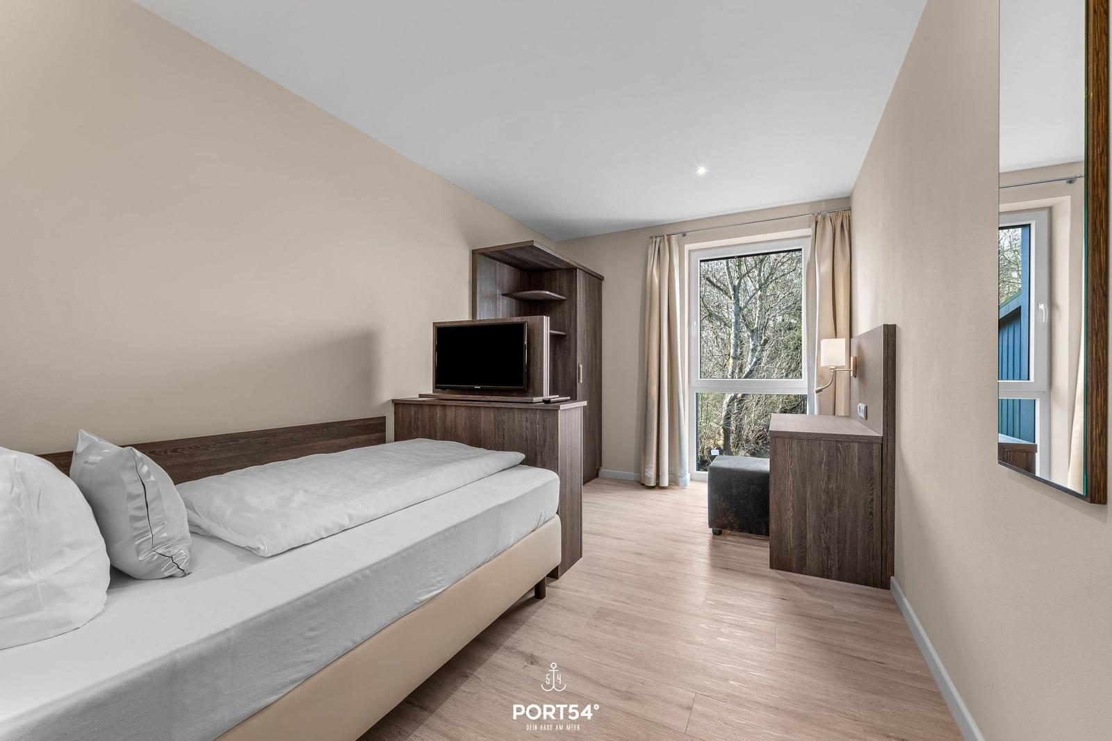 Schlafzimmer mit Bett, TV und Fensterblick auf Bäume.