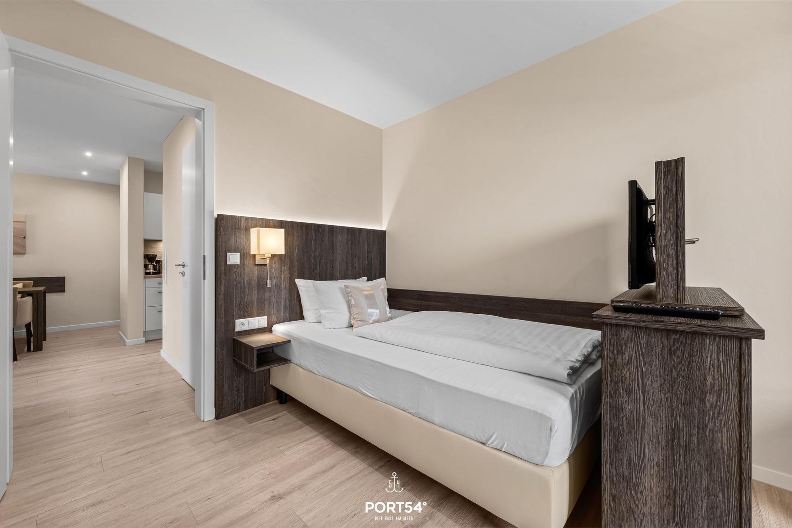 Schlafzimmer mit Bett, Nachttisch und Fernseher. Durchgang zur Küche und Esszone.