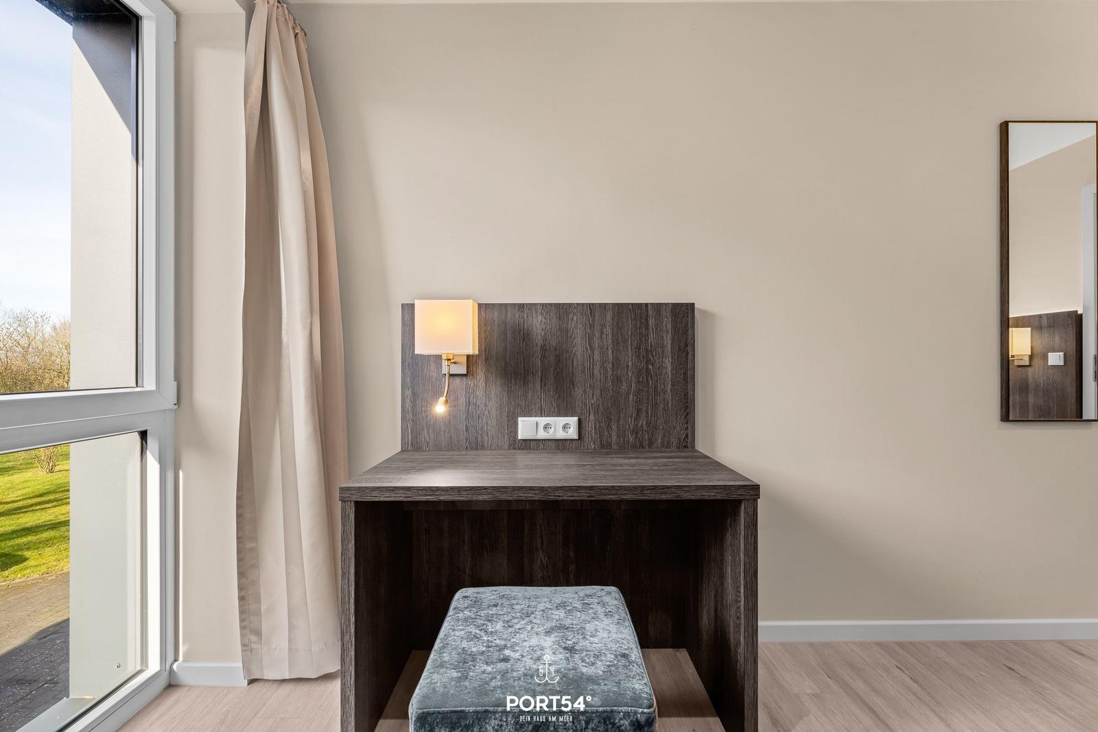 Schreibtisch mit Sitzbank, Wandlampe und Spiegel neben Fenster mit Blick auf Grünfläche.