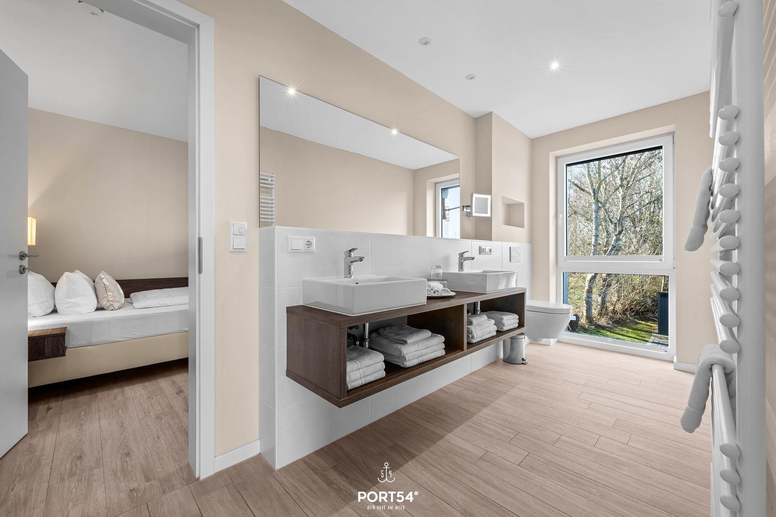 Badezimmer mit Doppelwaschbecken und Blick auf Garten. Schlafzimmer im Hintergrund.