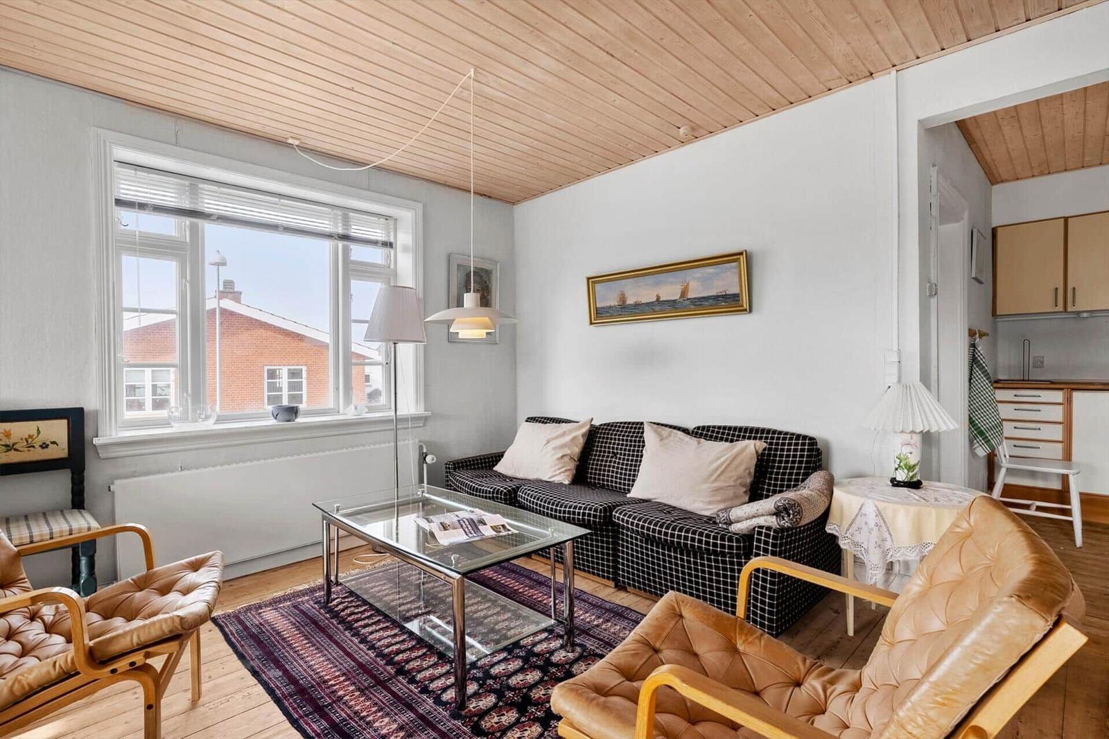 Living room with sofa, two leather chairs, and glass table. Wooden ceiling and window with view.