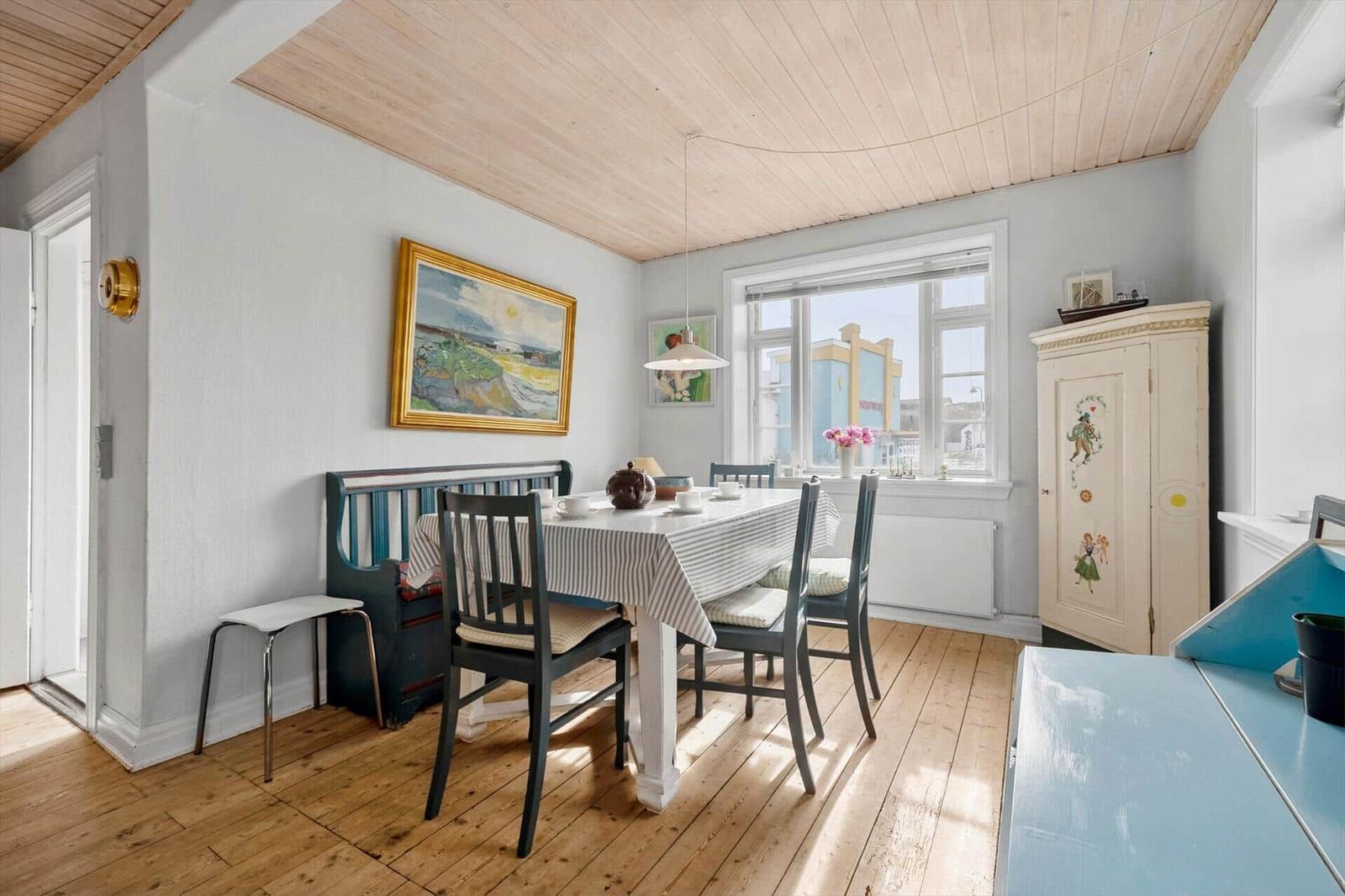 Dining room with table, chairs, and wooden floor. Window, paintings, and cabinet are visible.