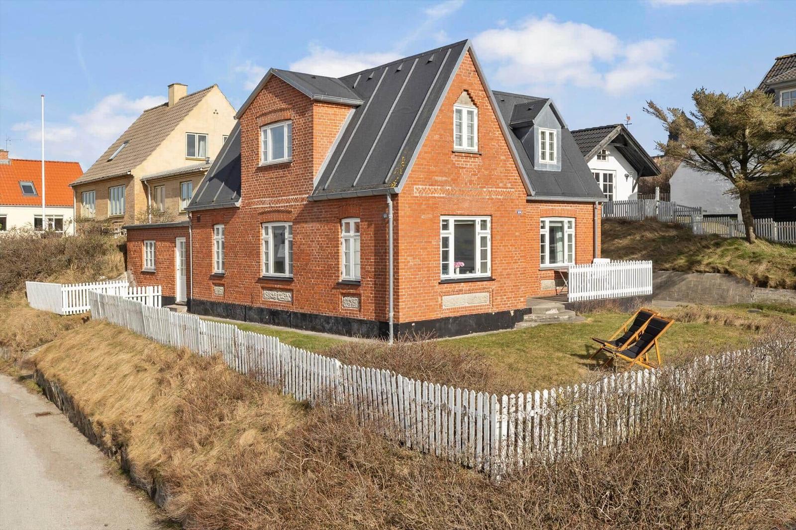 Red brick house with white windows and fence. Garden with sun lounger.