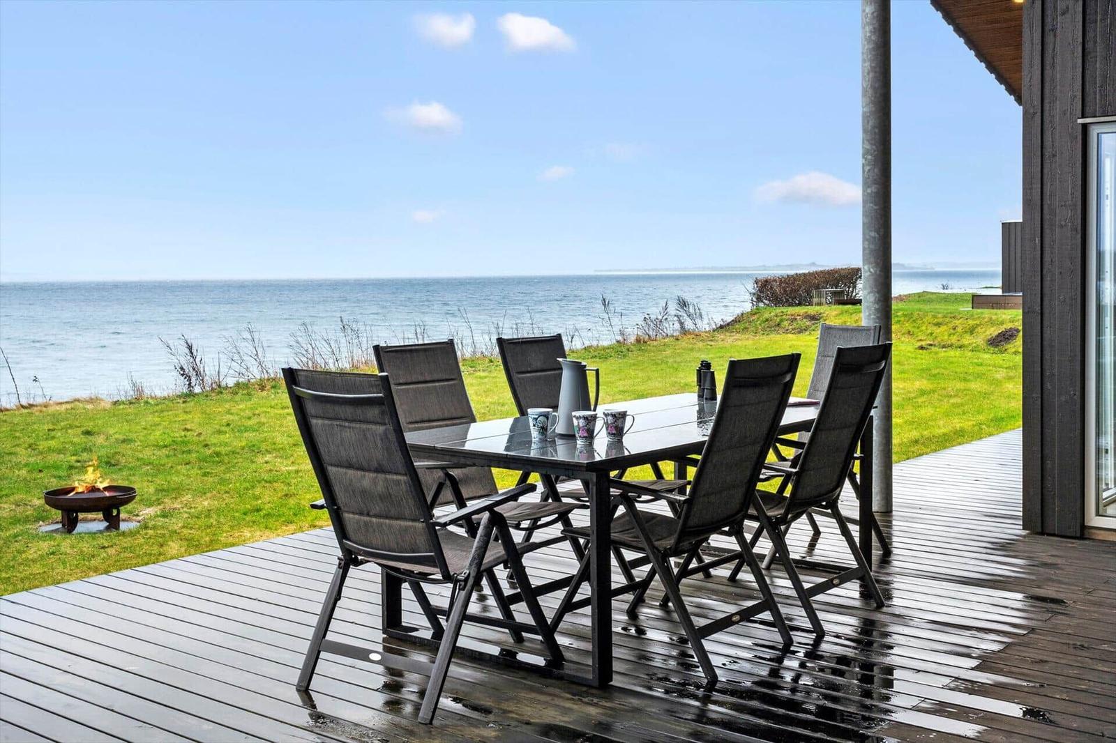 Deck with table and chairs, view of sea and grassy area.