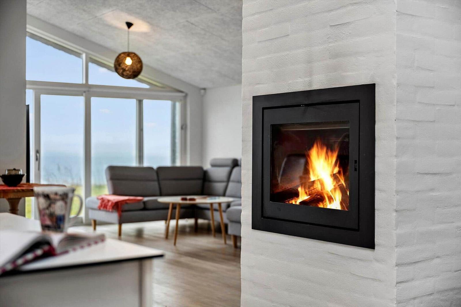 Fireplace with burning fire in white wall. Background shows living room with sofa and window.