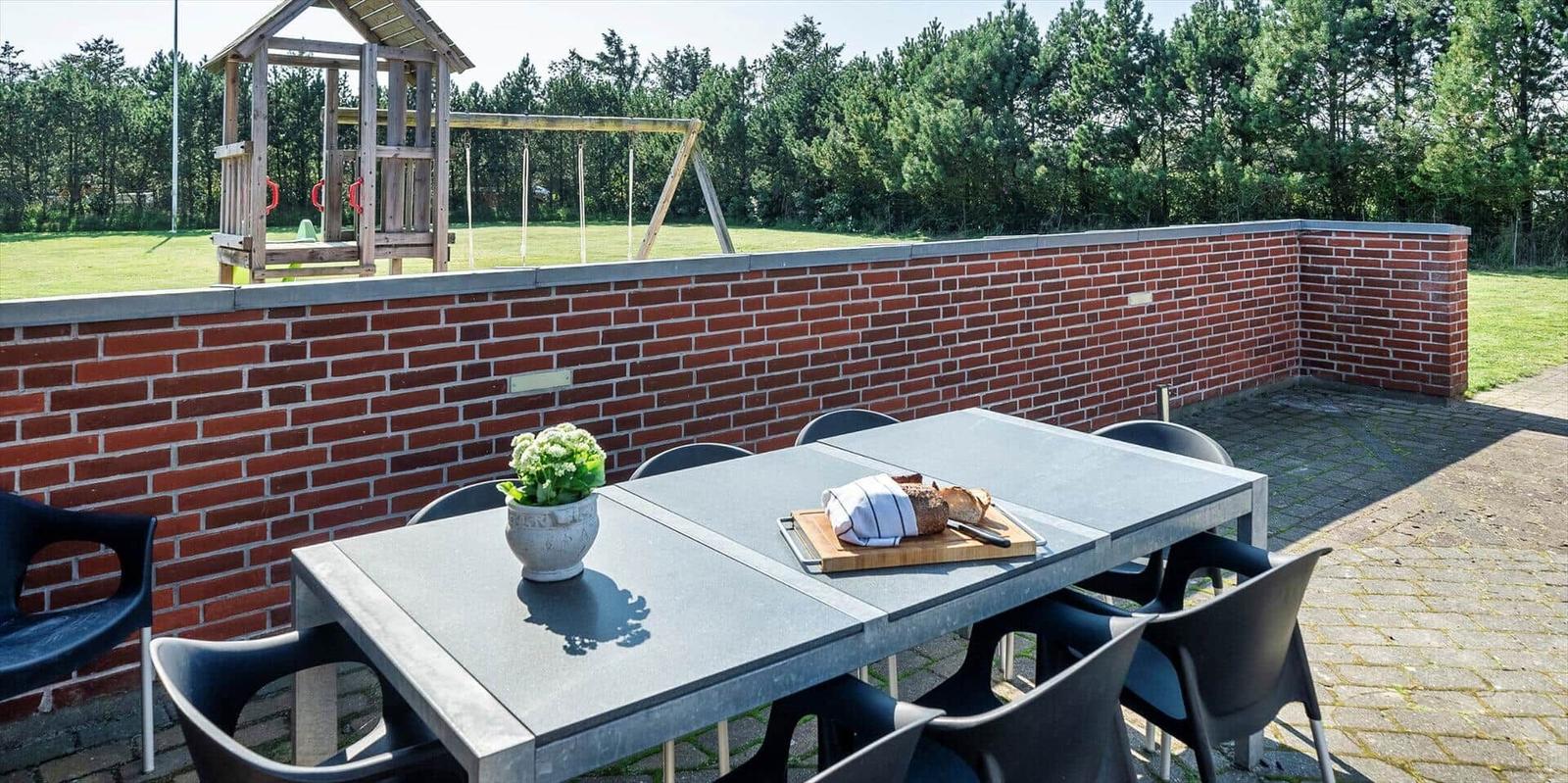 Terrace with table, chairs, plant, and bread. Backyard with playground and brick wall.