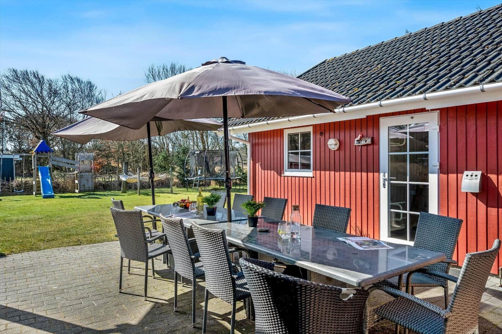 Red wooden house with terrace, dining table, and umbrella. Playground in the backyard.