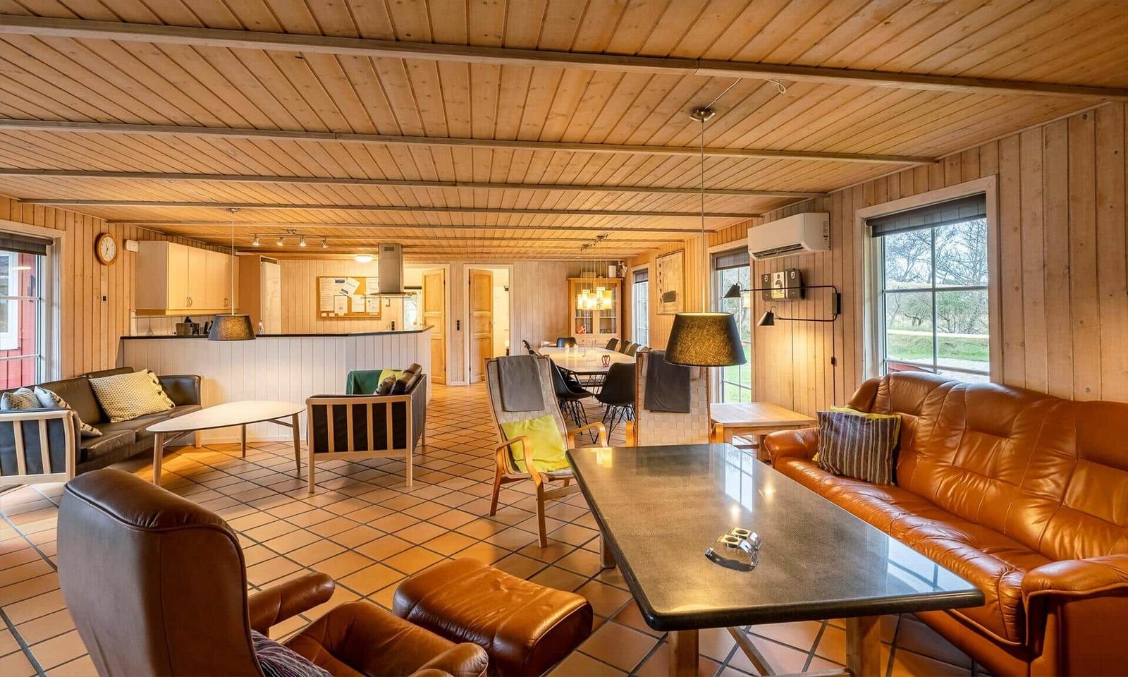 Living area with wooden walls, sofa, chairs, and dining table. Kitchen area in the background.