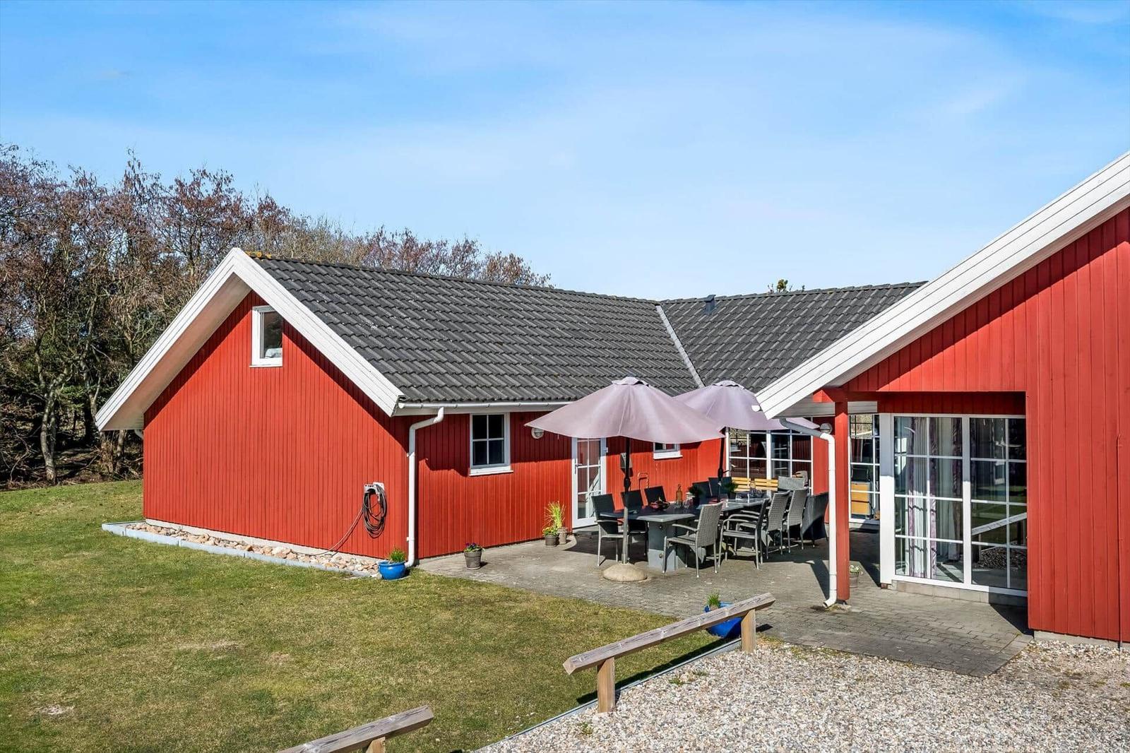 Red vacation house with terrace, table, chairs, and umbrellas. Lawn and trees in the background.