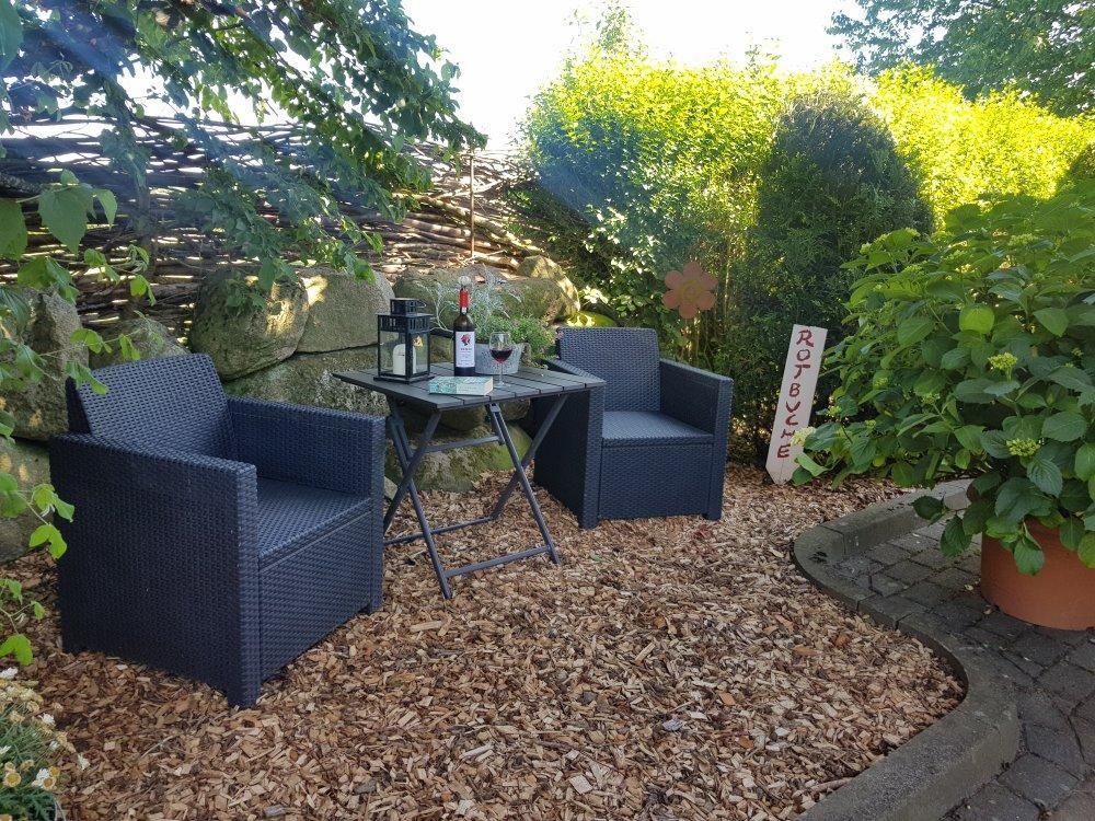 Garten mit zwei Sesseln, Tisch und Wein. Holzmulch, Pflanzen und Steinmauer.