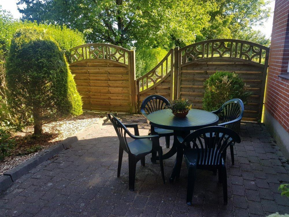Terrasse mit Tisch und Stühlen, Holztor und Beete im Hintergrund.