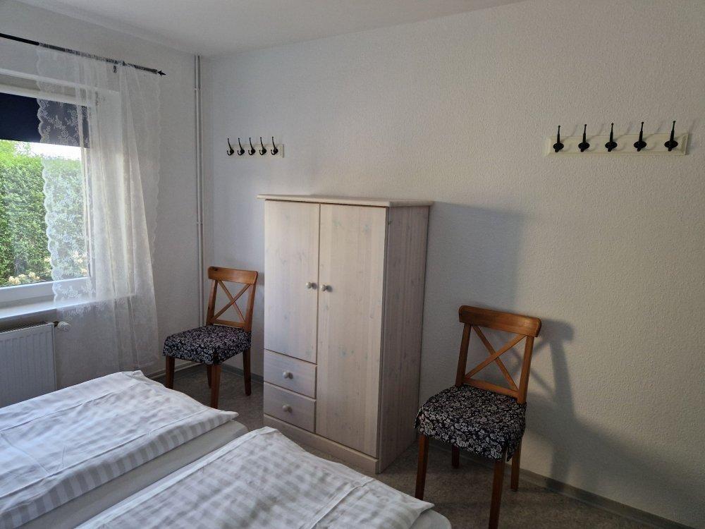 Zimmer mit Doppelbett, Kleiderschrank und zwei Stühlen. Fenster mit Vorhängen.