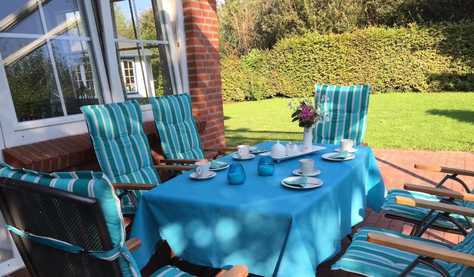 Terrasse mit gedecktem Tisch und Stühlen. Hintergrund: grüner Rasen und Hecke.