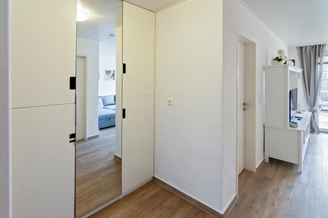 Flur mit Spiegeltüren, weißen Wänden und Holzboden. Blick in Wohnzimmer mit Sofa.