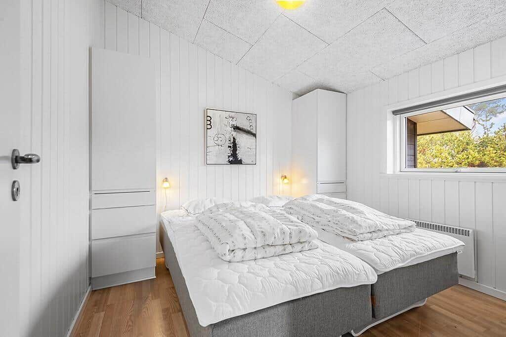 Schlafzimmer mit Doppelbett, weißen Schränken und Holzboden. Fenster zeigt Außenbereich.