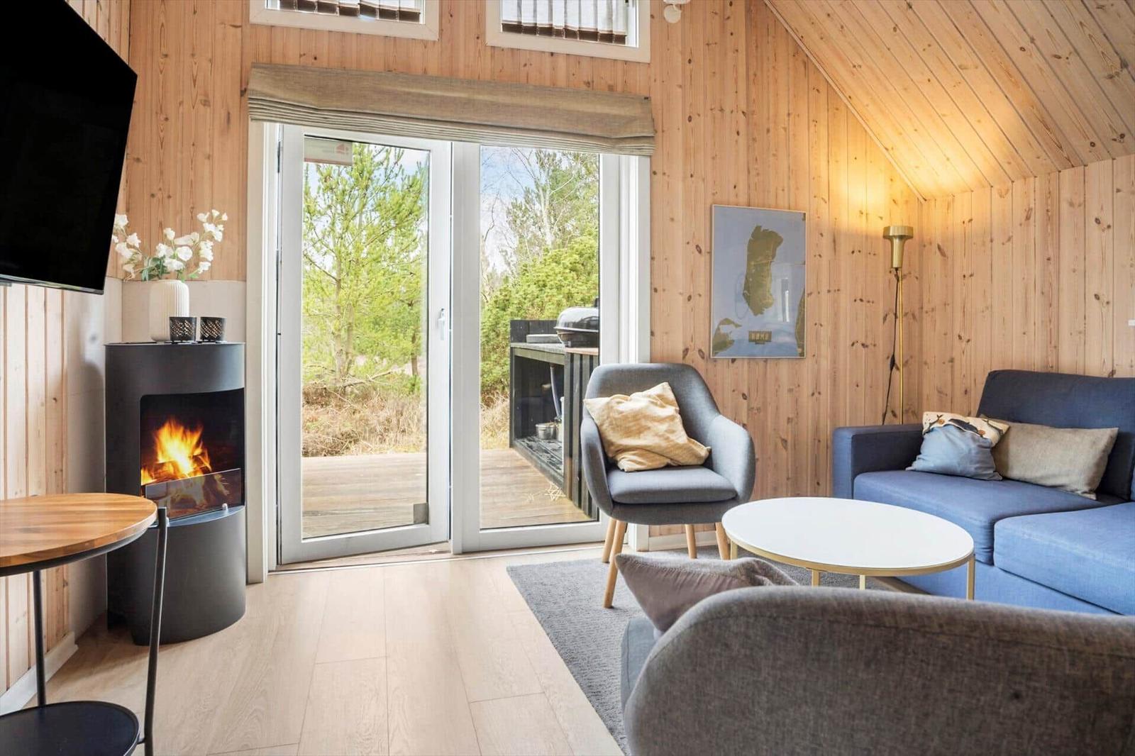 Wohnzimmer mit Kamin, Sofa und Sessel. Durchgang zur Terrasse mit Blick auf Wald.