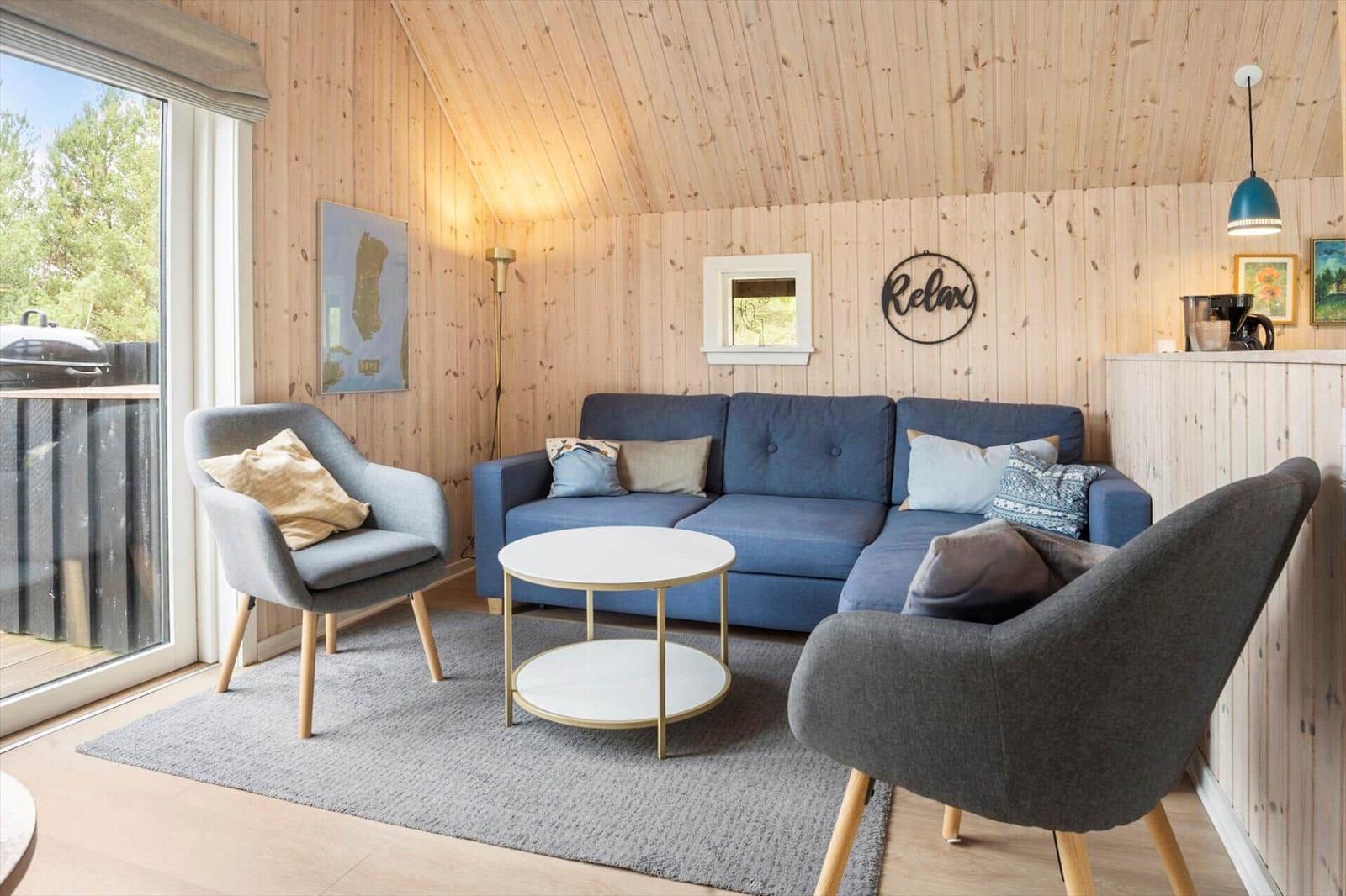 Wohnzimmer mit blauem Sofa, zwei Sesseln und runden Tisch. Holzwände und große Fenster.