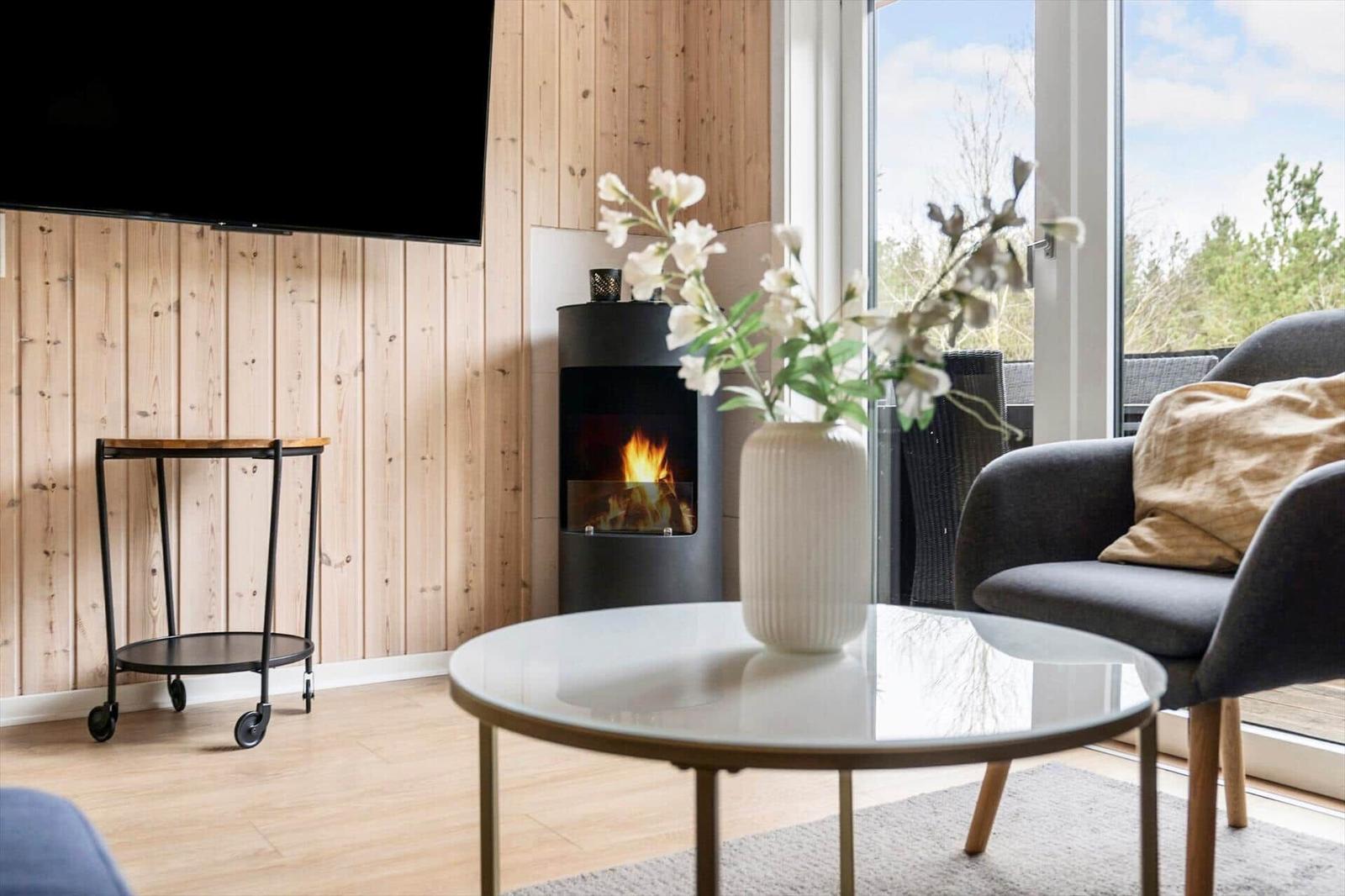 Wohnzimmer mit Kamin, Glas-Tisch und Sessel. Holzwand und großes Fenster.