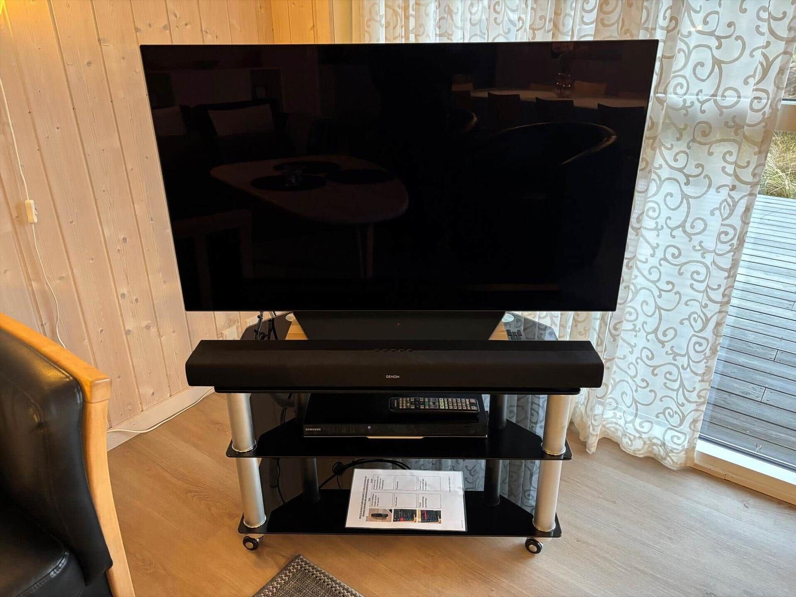 TV auf Stand mit Soundbar und Fernbedienung. Holzwand und Fenster mit Vorhang.