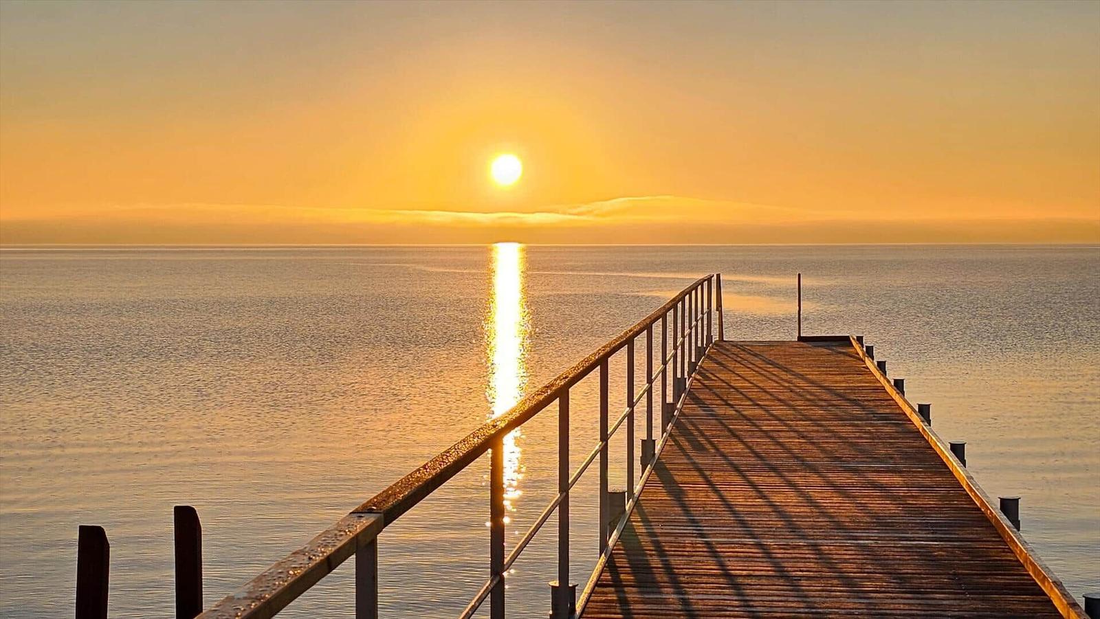 Holzsteg führt zum Horizont, Sonne spiegelt sich im Wasser.