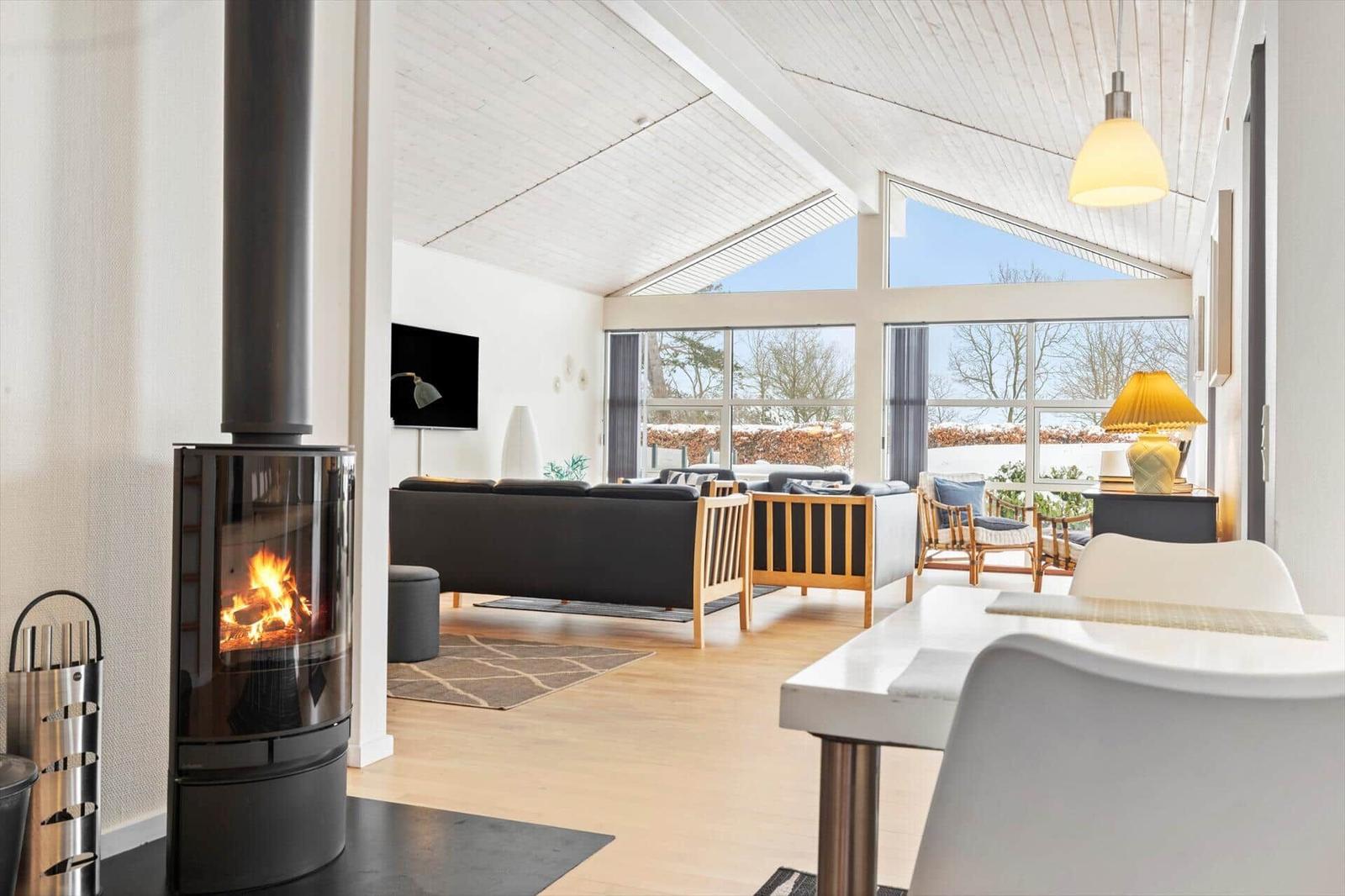 Wohnbereich mit Kamin, Sofa, Tisch und großen Fenstern. Holzboden und weißer Deckenrahmen.