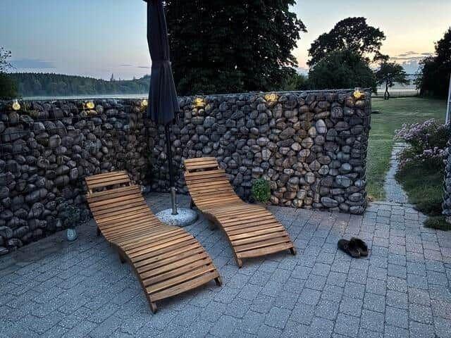 Zwei Holzliegestühle stehen auf einer gepflasterten Terrasse vor einer Steinmauer mit Beleuchtung.