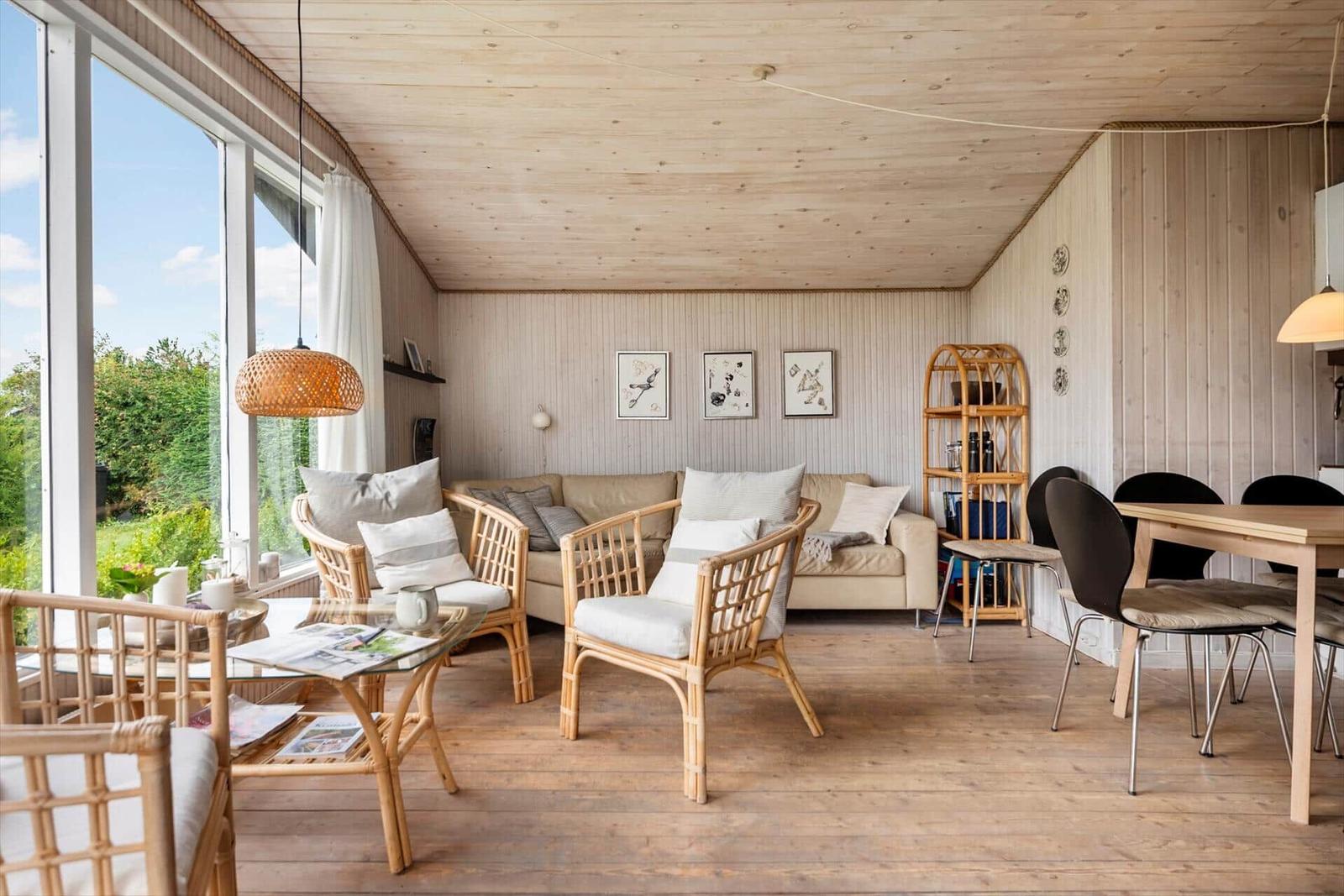 Wohnzimmer mit Holzdecke, Sitzgruppe, Tisch und Stühle. Große Fenster mit Blick auf Garten.