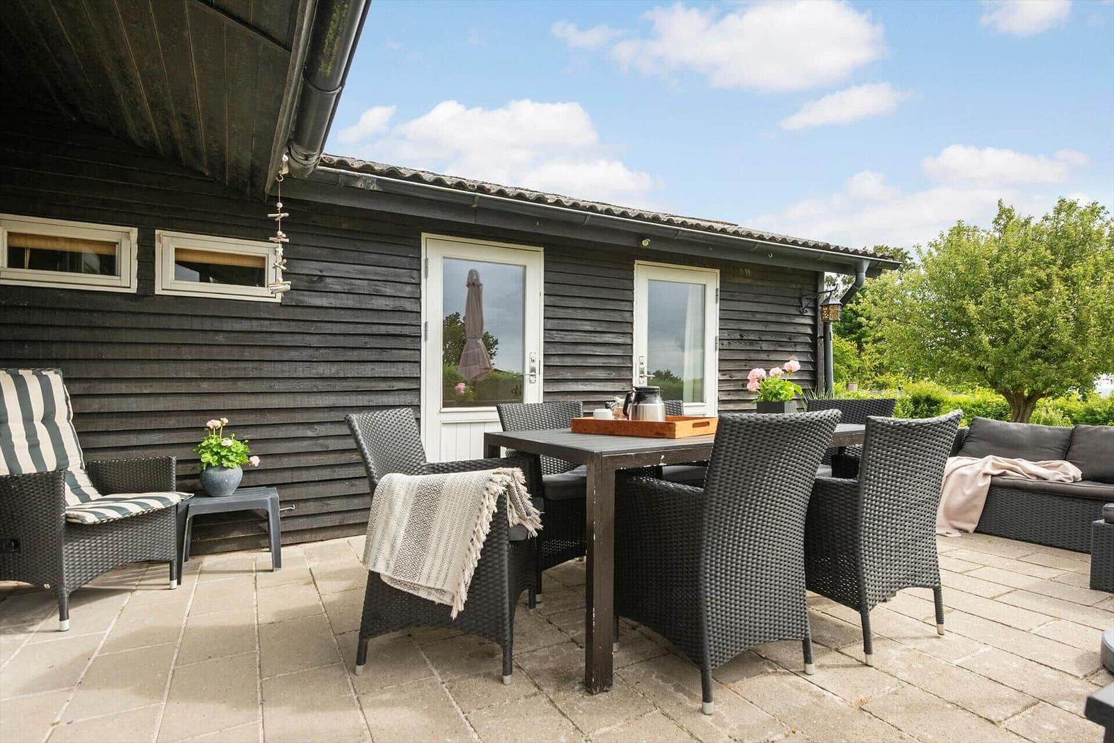 Terrasse mit Sitzgruppe, Tisch und Pflanzen vor einem dunklen Holzhaus.