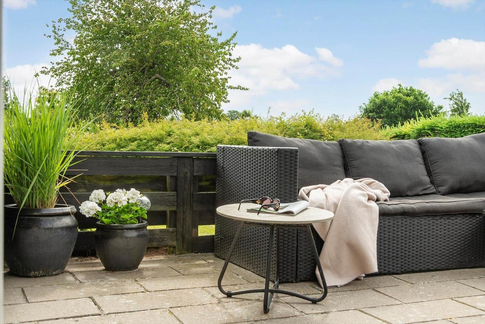 Terrasse mit Sitzgruppe, Tisch und Pflanzen. Hintergrund: grüne Hecke und Baum.