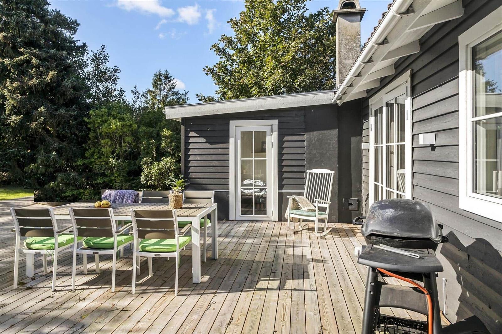 Terrasse mit Tisch, Stühlen und Grill. Hinterhof mit Bäumen und grauem Haus.