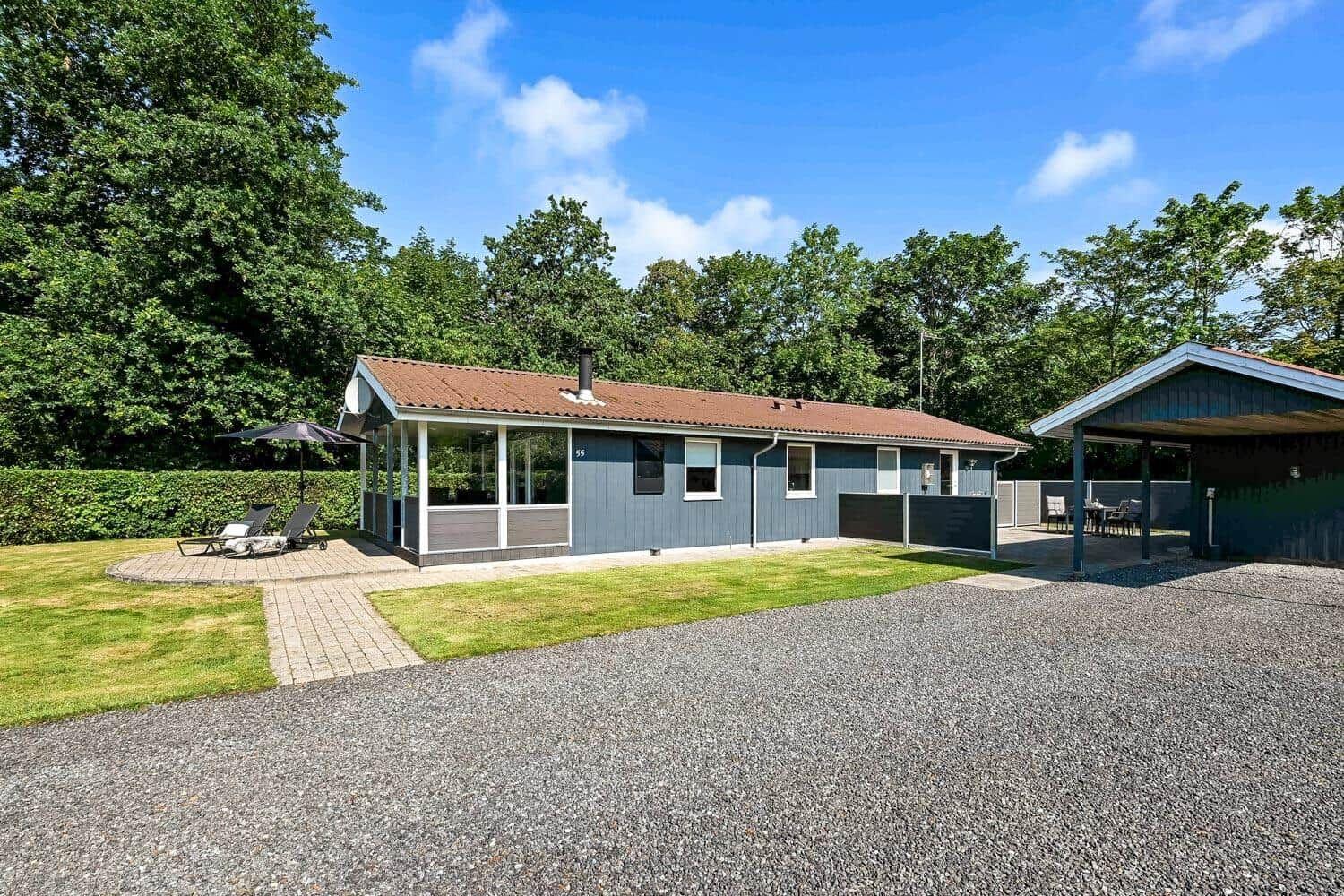 Ein graues Ferienhaus mit Terrasse, Liegestühlen und Grillplatz. Grün umgeben mit Wald.