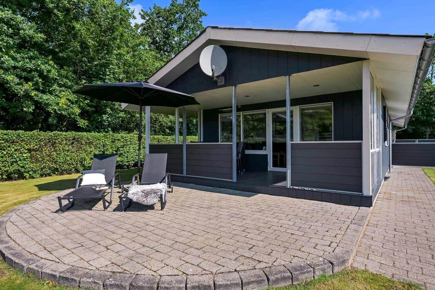 Haus mit überdachter Terrasse, Liegestühle und Sonnenschirm. Grün umgeben.