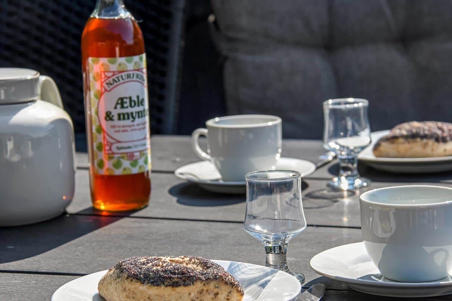 Außenbereich mit Tisch, Tassen, Gläsern und Gebäck. Eine Flasche mit Getränk steht daneben.