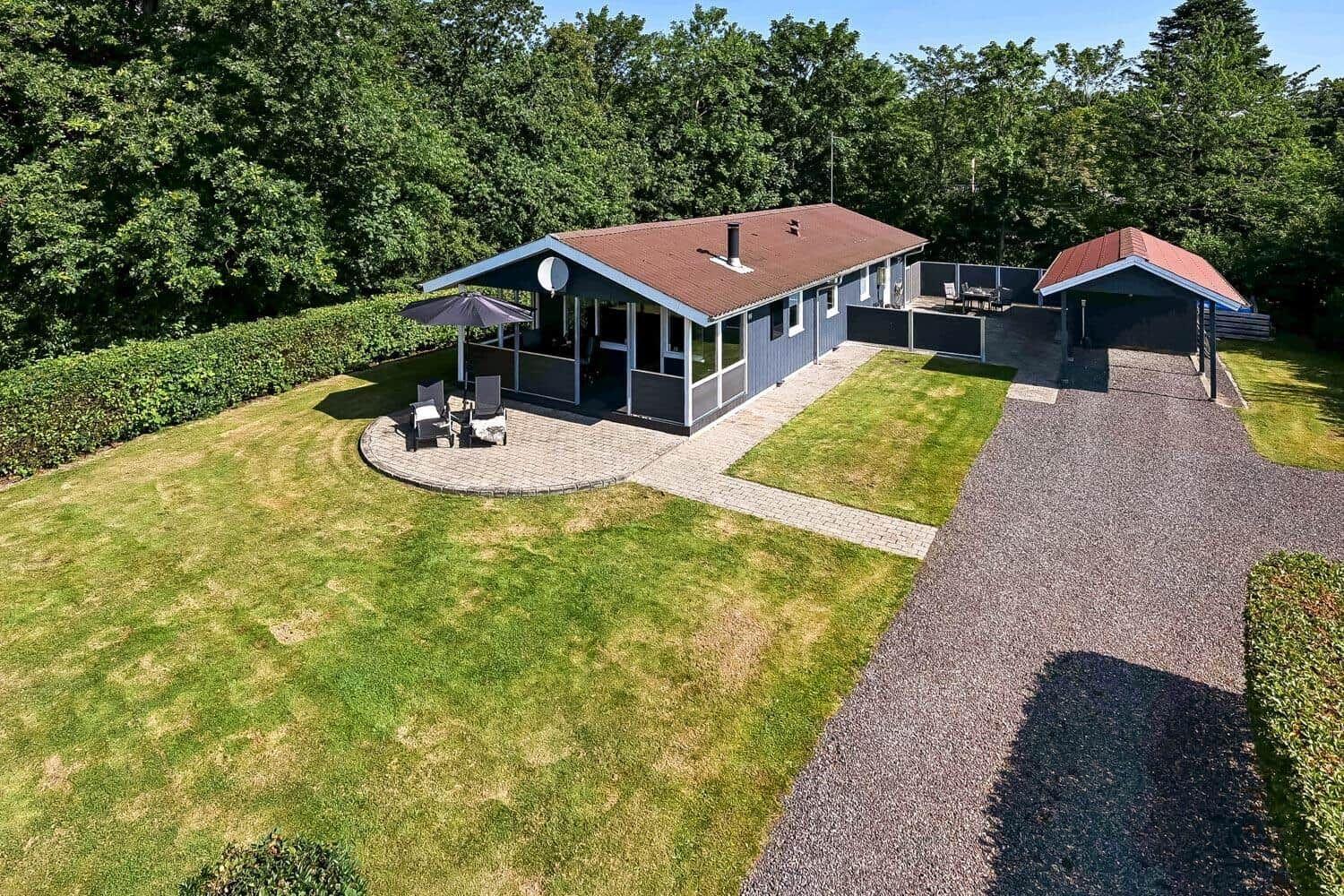 Ein Ferienhaus mit Terrasse, Grillplatz und Rasenfläche. Umgeben von Bäumen und einem Kiesweg.