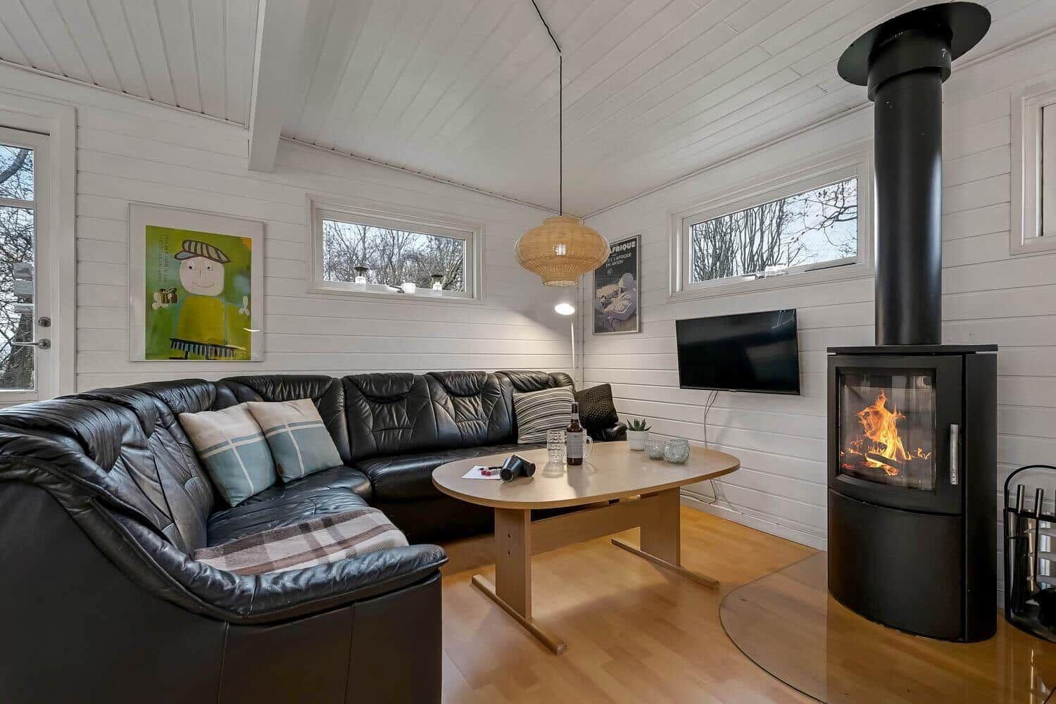 Wohnzimmer mit schwarzer Ledercouch, Kamin und Holztisch. An den Wänden hängen Bilder.