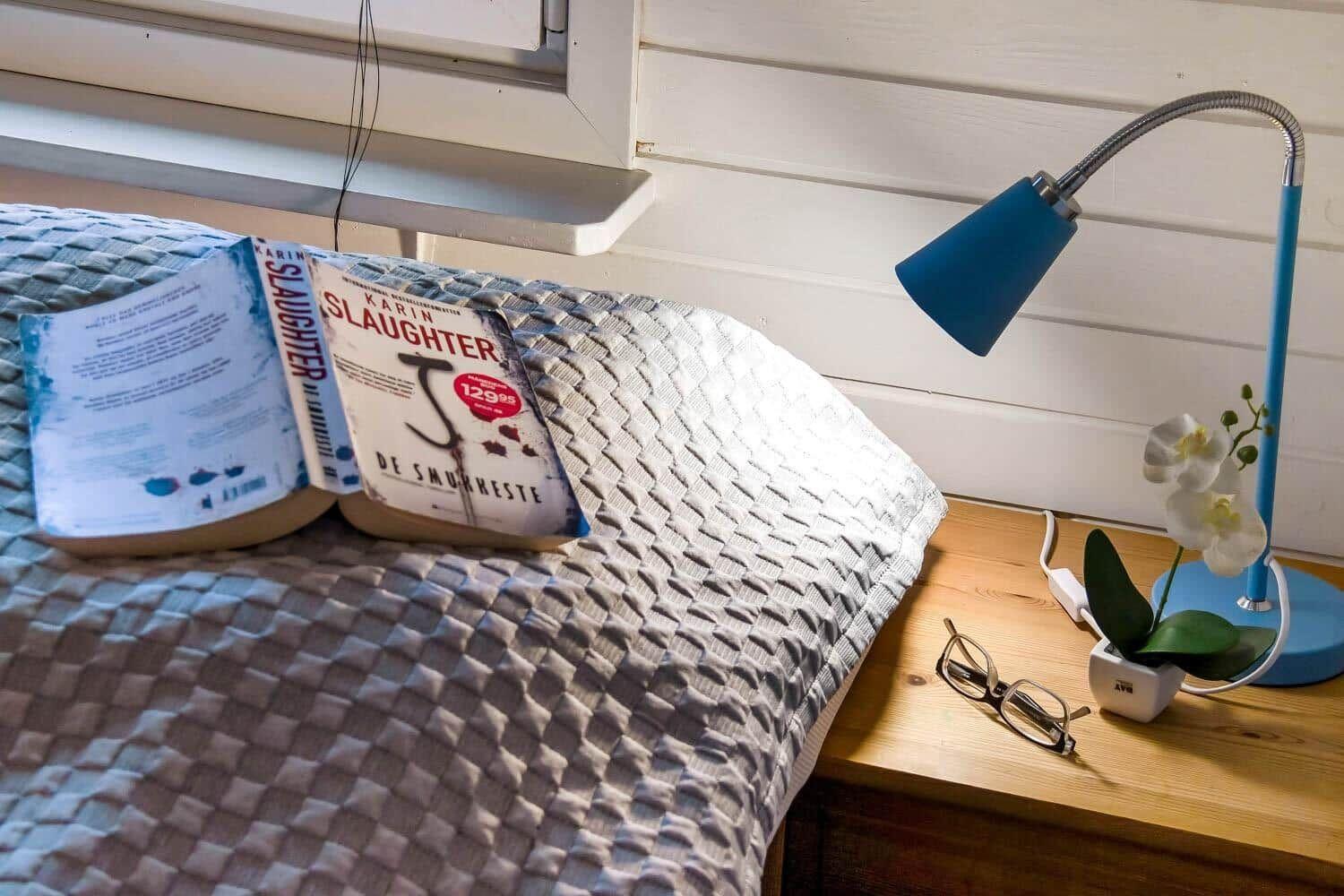 Ein Bett mit einer Decke, einem Buch und einer Lampe neben einem Nachttisch.
