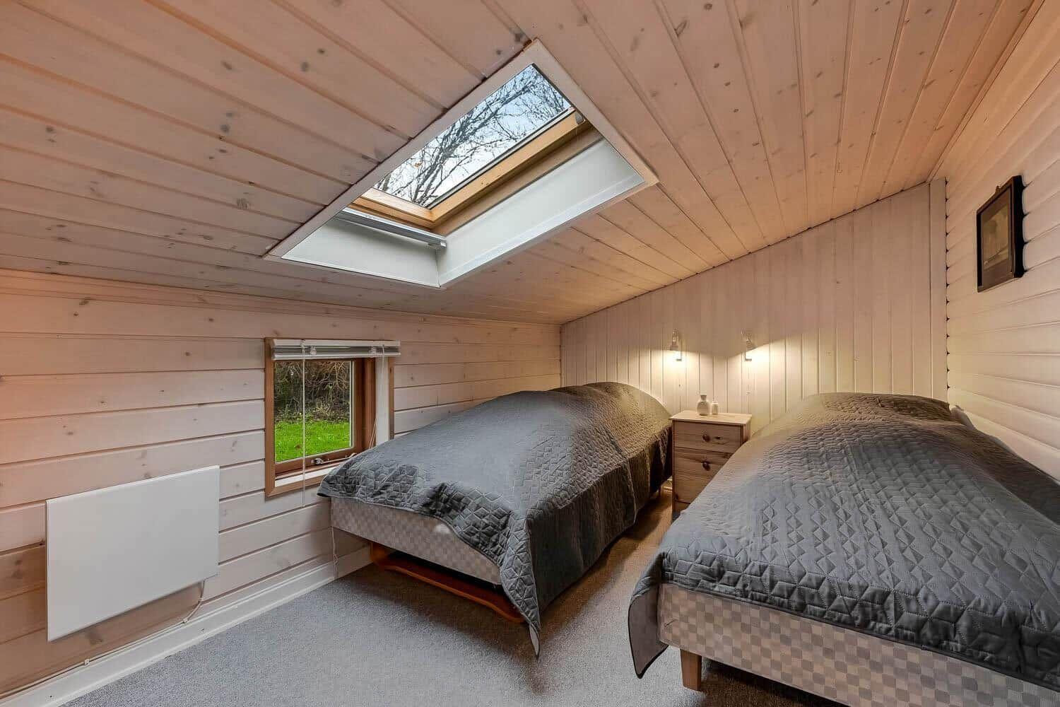 Zwei Betten in einem Zimmer mit Holzwand und Dachfenster. Kleines Fenster und Nachttisch.