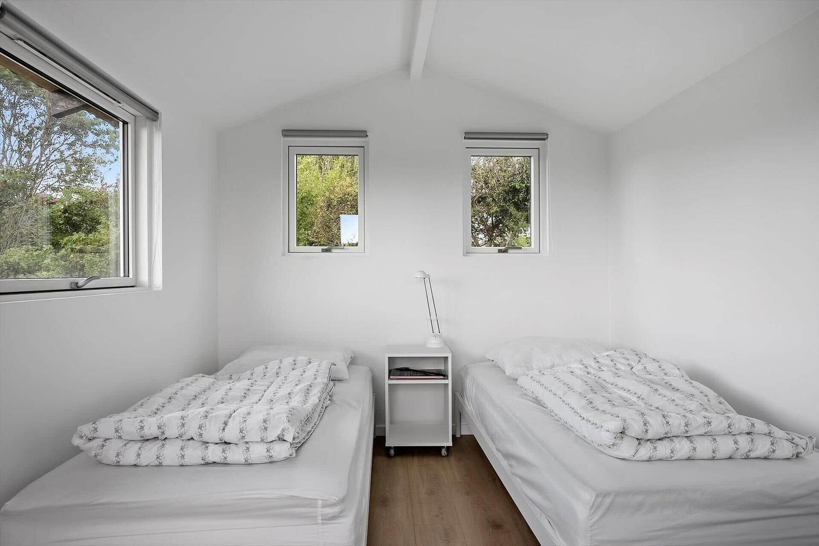 Zwei Betten mit weißen Laken und gemusterten Decken. Drei Fenster mit Blick auf Grün.