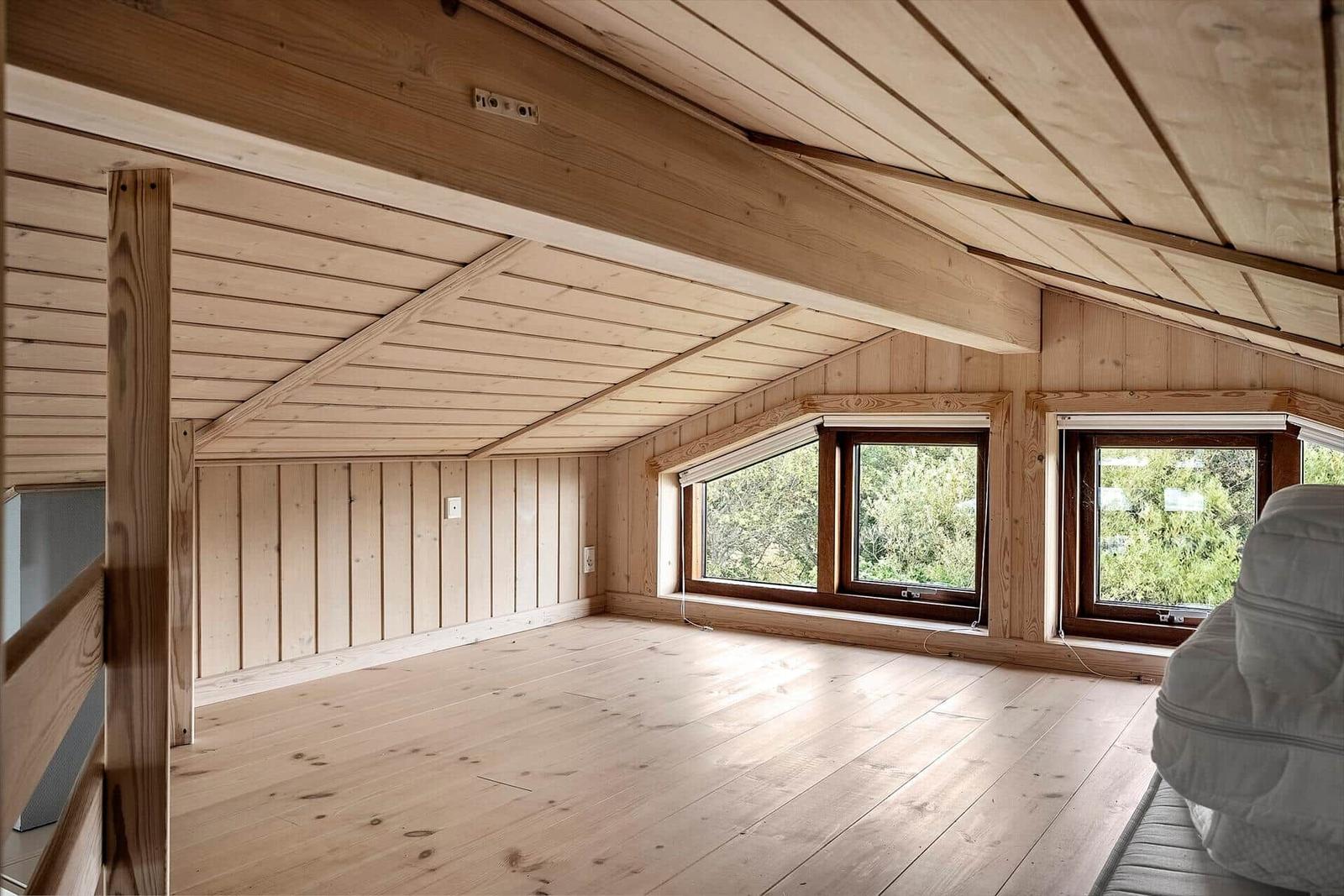 Wohnraum mit Holzwand, Holzdecke und Fenstern. Blick auf grüne Bäume.